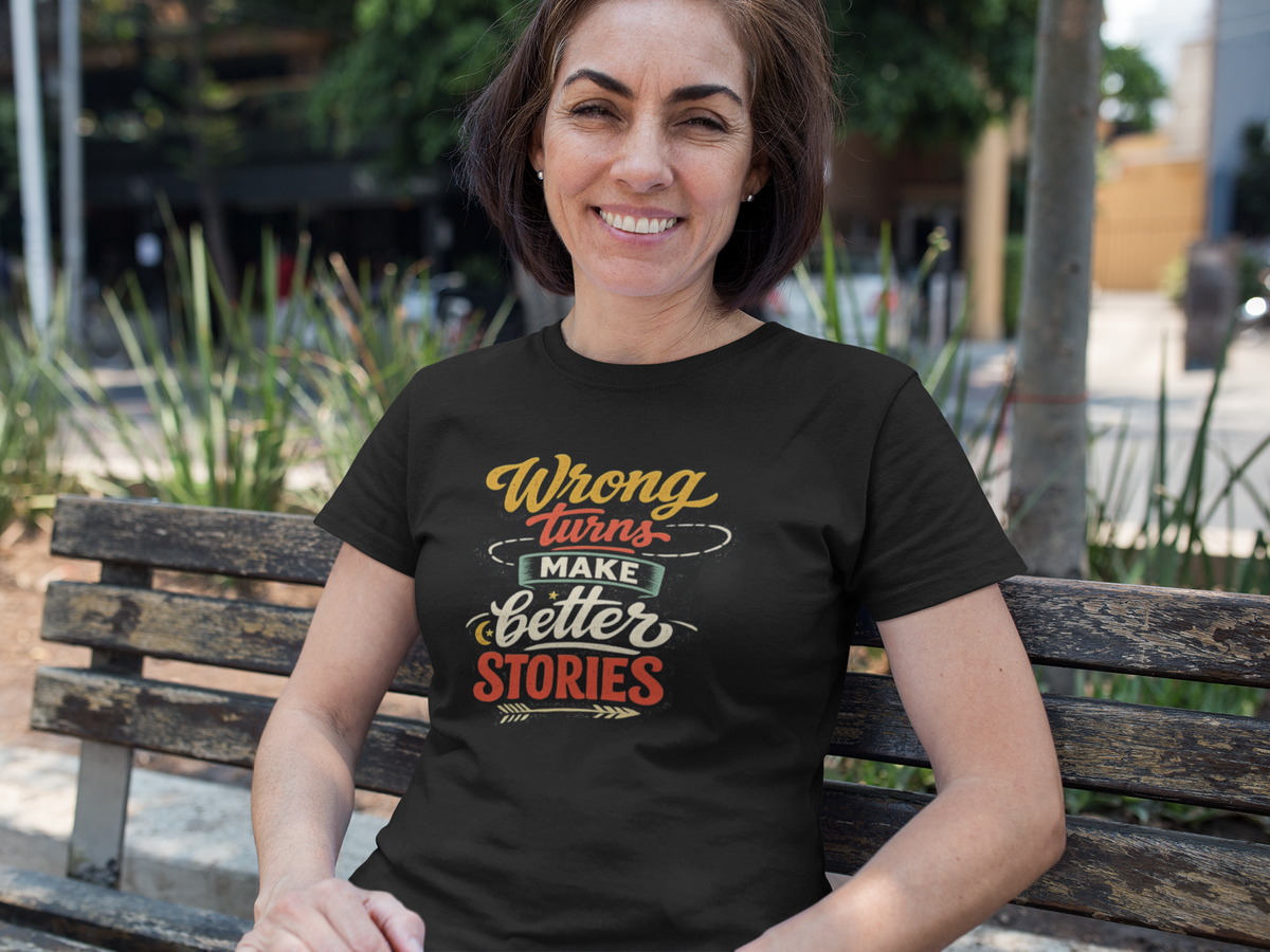 A woman sitting on a wooden bench outdoors wearing a black graphic t-shirt with the text 'Wrong Turns Make Better Stories' in yellow, white, and red lettering.