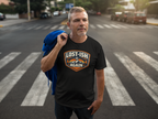 A man standing on a city crosswalk wearing a black graphic t-shirt with a 'Lost-Ish Again' patch design, holding a blue jacket over his shoulder.