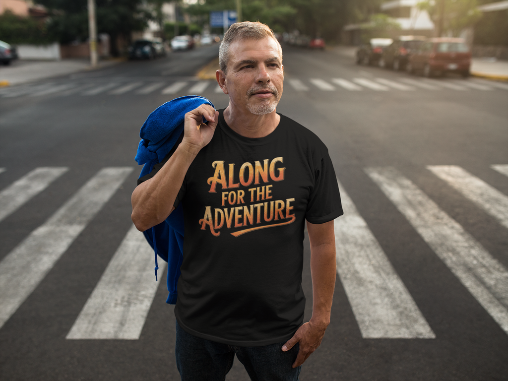 A man standing on a city crosswalk wearing a black graphic t-shirt with the text 'ALONG FOR THE ADVENTURE' in a stylized orange and gold font, and carrying a blue backpack over his shoulder.