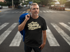 A man standing on a city crosswalk wearing a black graphic t-shirt with 'Always Rolling Forward' printed in a gold-colored cursive font. He has a blue jacket slung over his shoulder, with a blurred street background.