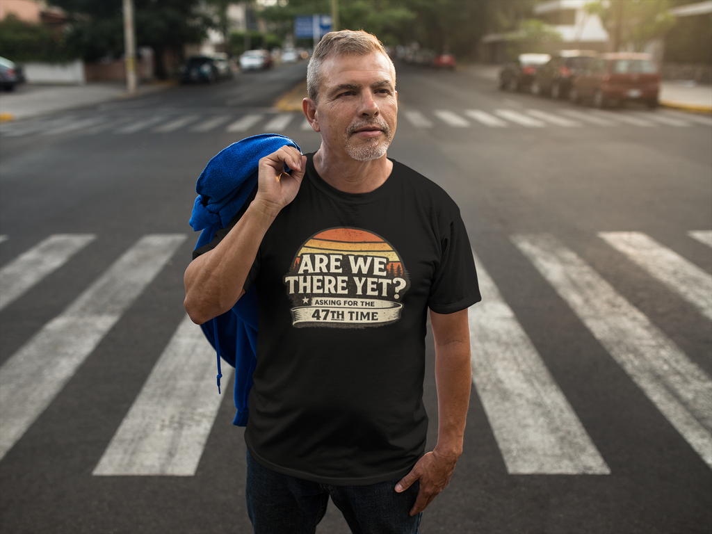 A man standing on a city crosswalk wearing a black graphic t-shirt with a retro-style orange and white design that reads 'ARE WE THERE YET? Asking for the 47TH TIME'. He has a blue jacket slung over his shoulder, with a blurred street background.