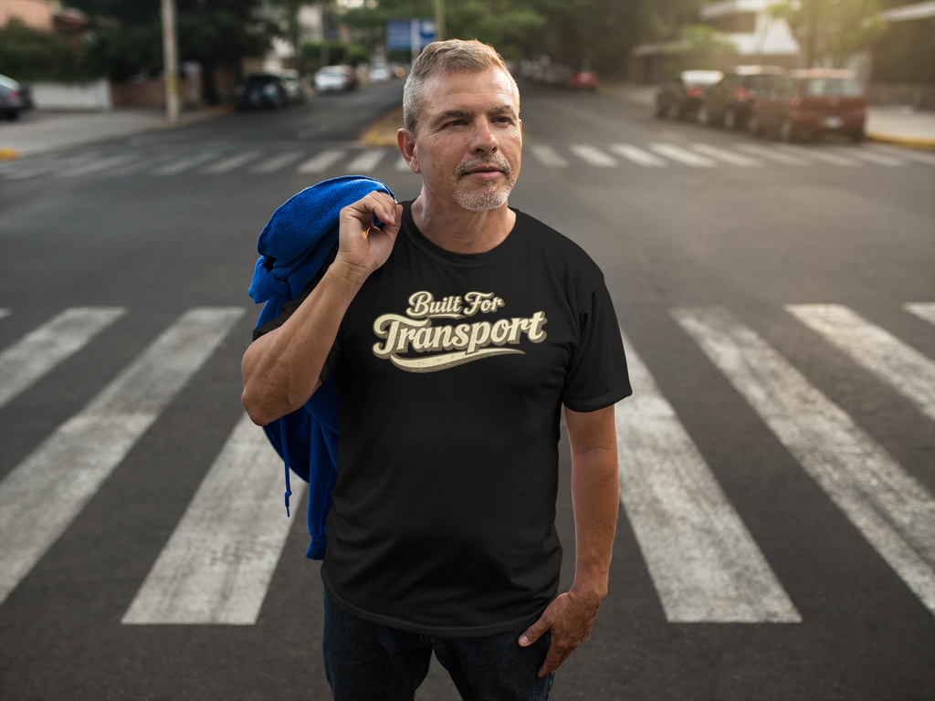 A man standing on a city crosswalk wearing a black t-shirt with a gold graphic that reads 'Built For Transport'. He has a blue jacket slung over his shoulder, with a blurred street background.