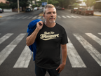 A man standing on a city crosswalk wearing a black t-shirt with a gold graphic that reads 'Built For Transport'. He has a blue jacket slung over his shoulder, with a blurred street background.