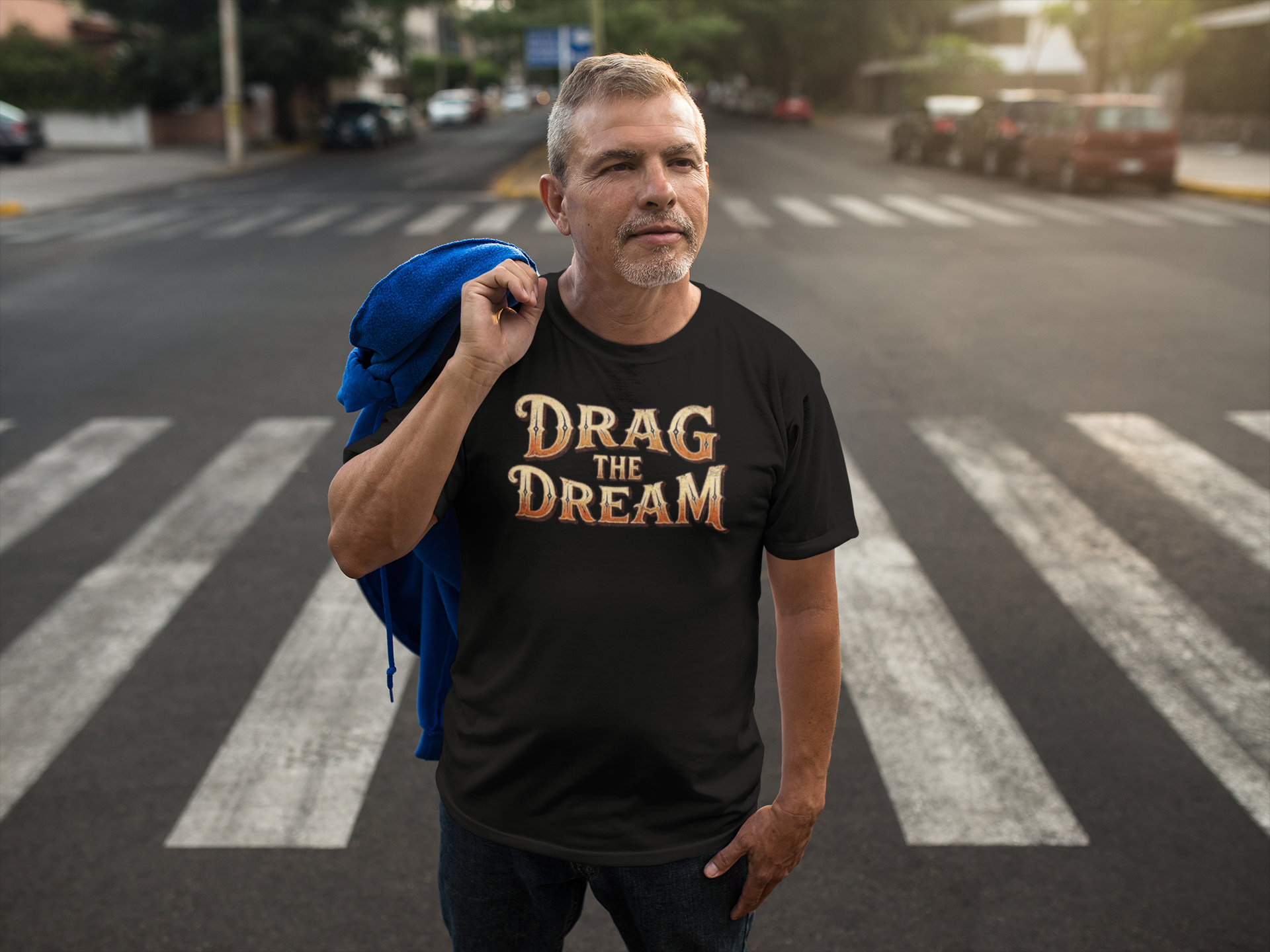 A man standing on a city crosswalk wearing a black graphic t-shirt with the text 'DRAG THE DREAM' in a gold and orange gradient. He has a blue jacket draped over his shoulder, with a blurred street background.