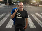 A man standing on a city crosswalk wearing a black t-shirt with the text 'Eat. Tow Sleep'