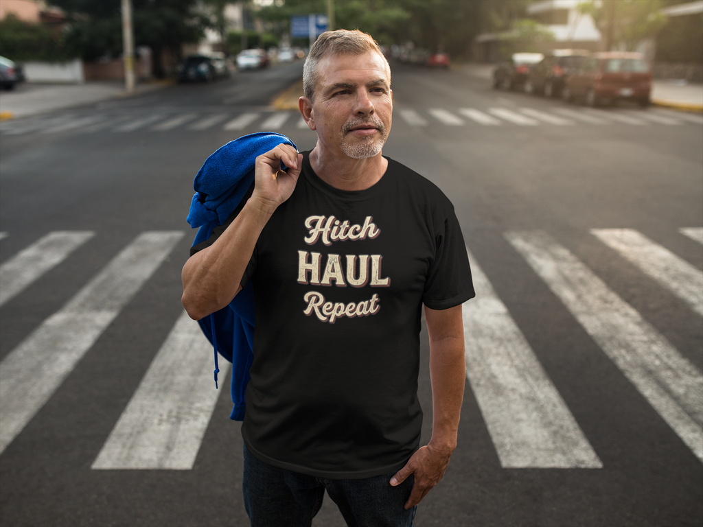 A man standing on a crosswalk wearing a black graphic t-shirt that reads 'Hitch HAUL Repeat' in beige lettering, with a blue jacket slung over his shoulder, set against a blurred street background.