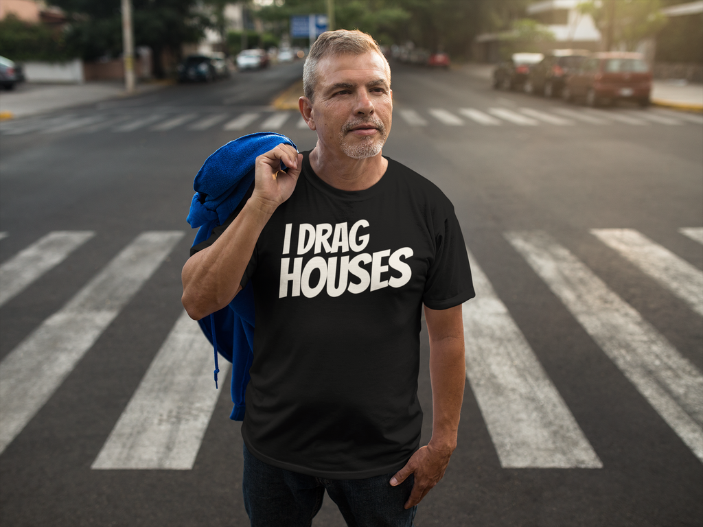 A man standing on a city crosswalk wearing a black graphic t-shirt with the text 'I DRAG HOUSES' in bold white letters, with a blue jacket slouched over his shoulder.