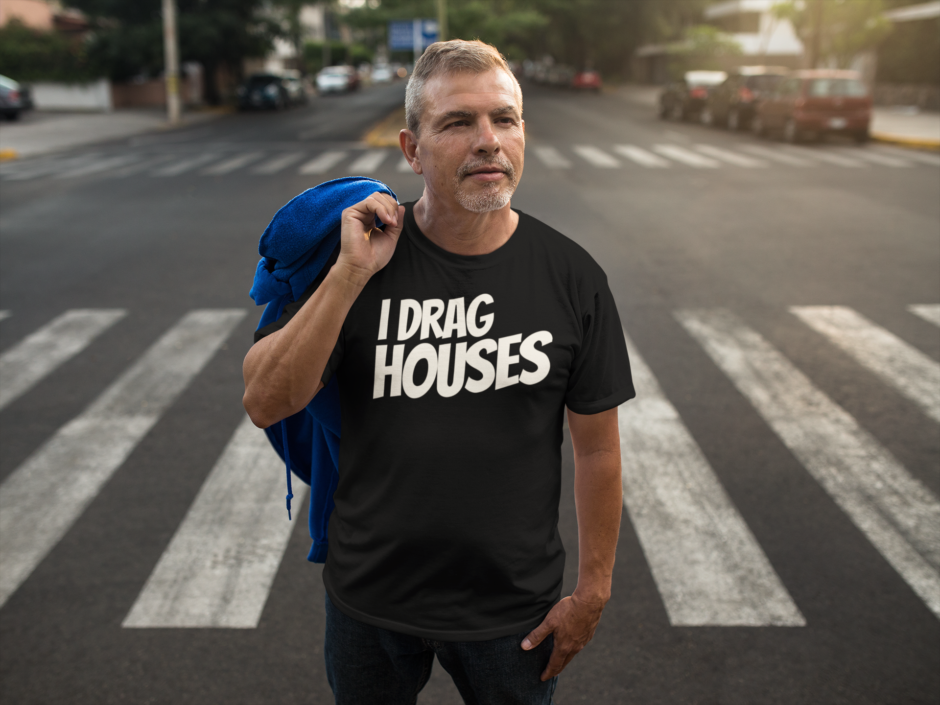 A man standing on a city crosswalk wearing a black graphic t-shirt with the text 'I DRAG HOUSES' in bold white letters, with a blue jacket slouched over his shoulder.