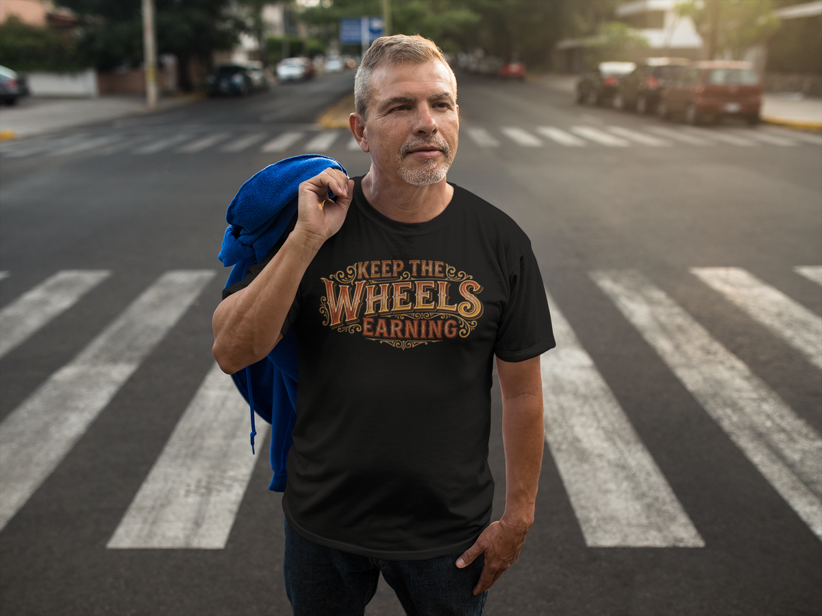 A man standing on a city crosswalk wearing a black graphic t-shirt with the text 'KEEP THE WHEELS EARNING' in a distressed gold and brown font. He has a blue jacket draped over his shoulder, with a blurred street background.