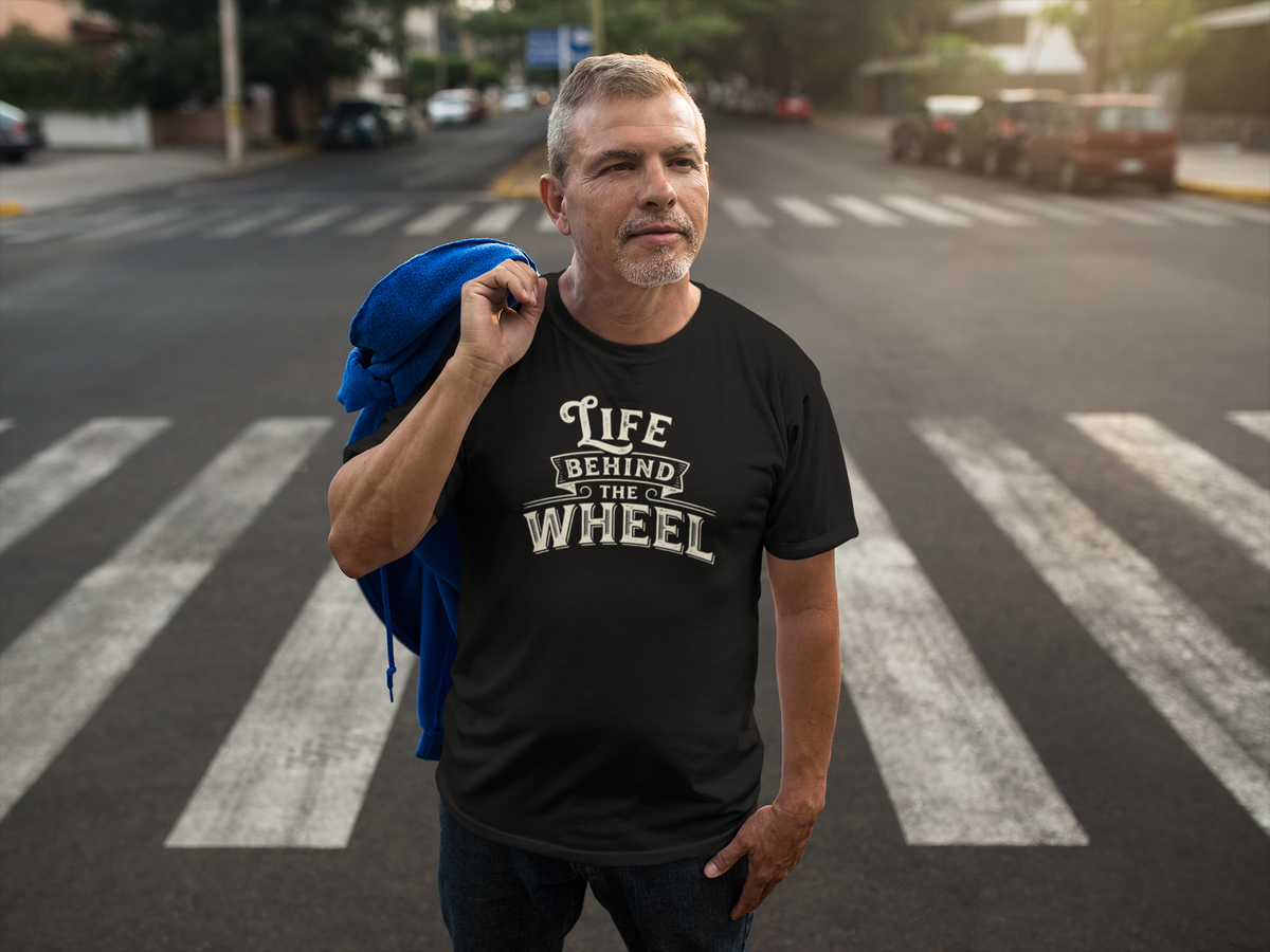 A man standing on a city crosswalk wearing a black graphic t-shirt with the text 'Life Behind the Wheel' in a distressed white font, holding a blue jacket over his shoulder.