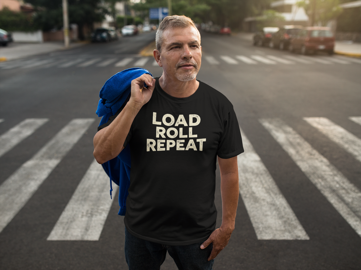 A man standing on a city crosswalk wearing a black t-shirt with the text 'LOAD ROLL REPEAT' in beige, with a blue backpack slung over his shoulder.