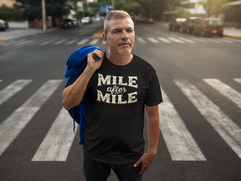 A man standing on a city crosswalk wearing a black t-shirt with 'MILE after MILE' printed in beige, holding a blue jacket over his shoulder.