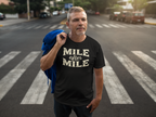 A man standing on a city crosswalk wearing a black t-shirt with 'MILE after MILE' printed in beige, holding a blue jacket over his shoulder.