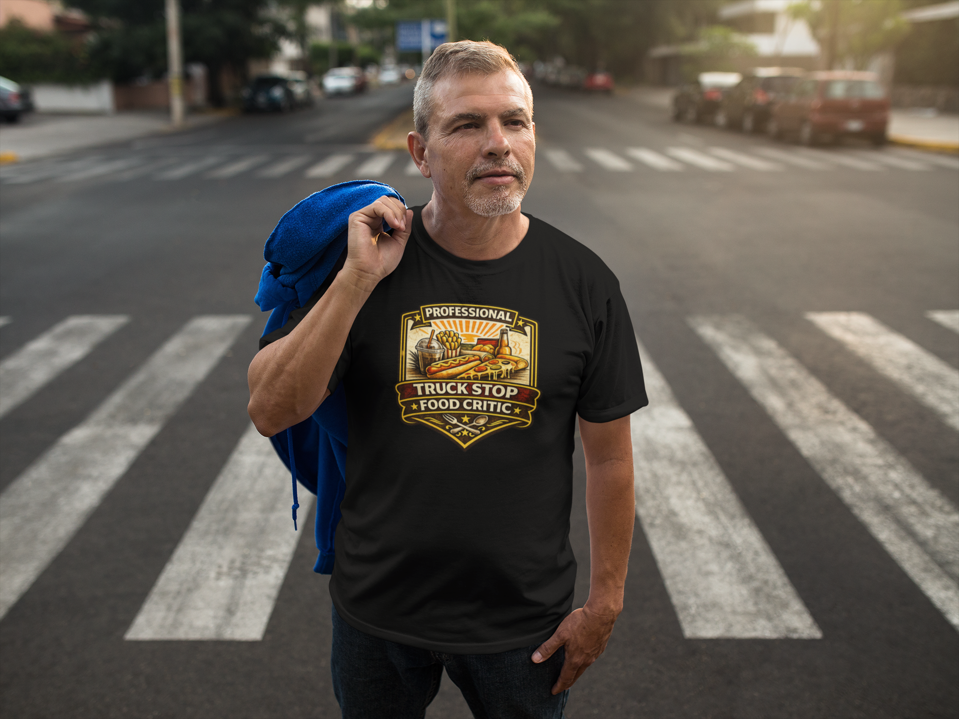 A man standing on a city crosswalk wearing a black graphic t-shirt with a 'Professional Truck Stop Food Critic' badge design. He has a blue jacket draped over his shoulder, with a blurred street background.