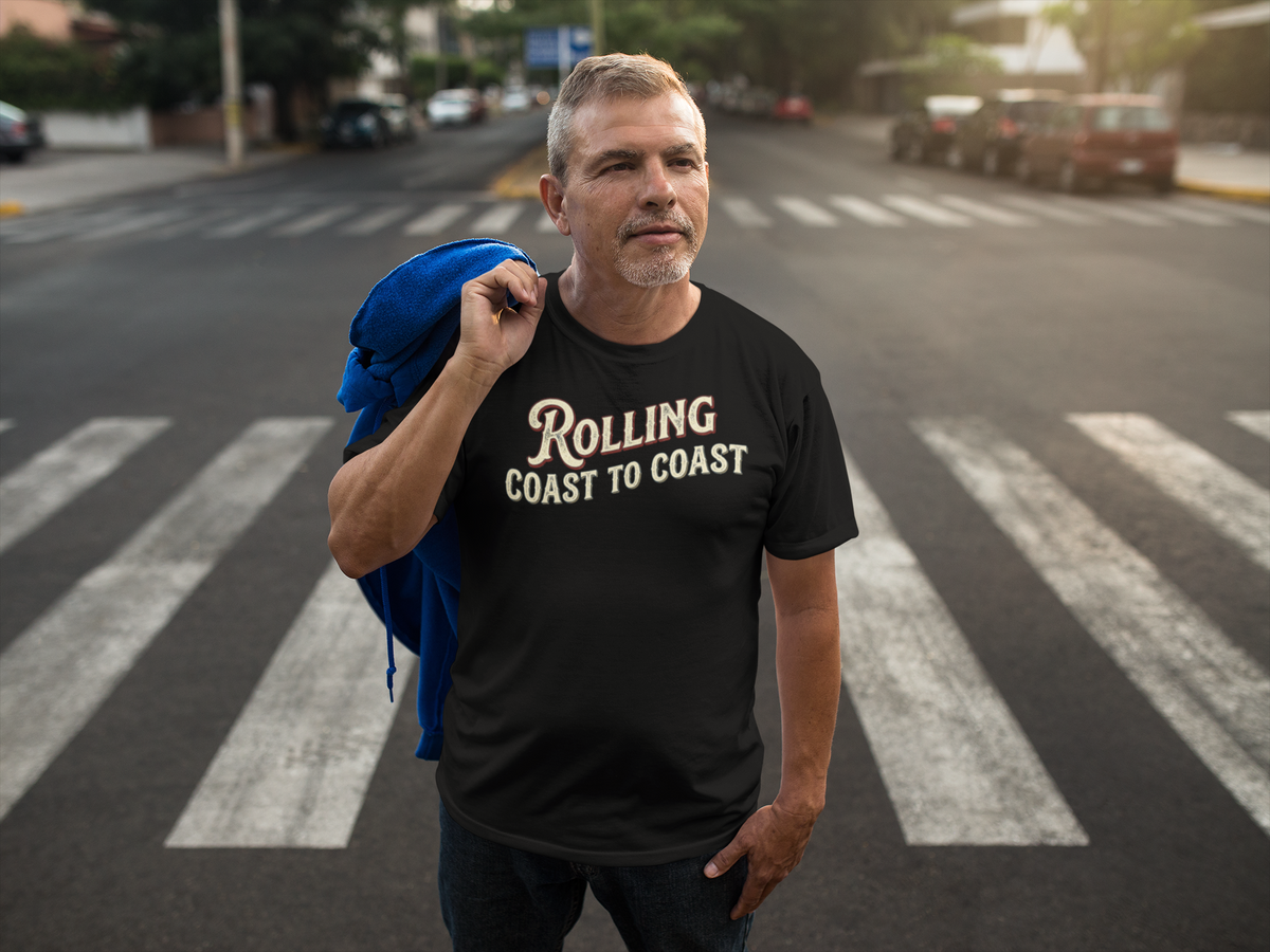 A man standing on a city crosswalk wearing a black graphic t-shirt with 'INTERSTATE RUNNER' printed in a stylized beige and gold font. He has a blue jacket draped over his shoulder, with a blurred street background.
