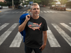 A man standing on a city crosswalk wearing a black t-shirt with a graphic of a USA map filled with the American flag and the text 'Rolling Through The Red White & Blue The Riverstone Life'. He is holding a blue jacket over his shoulder.