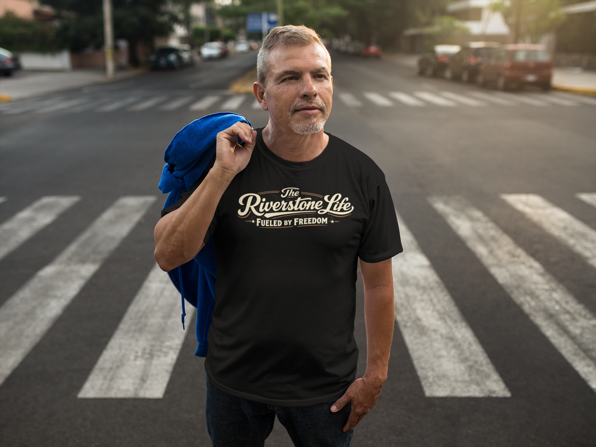 A man standing on a city crosswalk wearing a black graphic t-shirt with 'The Riverstone Life Fueled By Freedom' printed in a stylized beige and gold font. He has a blue jacket draped over his shoulder, with a blurred street background.