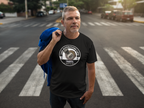 A man standing on a city crosswalk wearing a black t-shirt with a circular 'The Riverstone Life Original' graphic, holding a blue jacket over his shoulder.