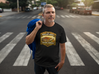 A man standing on a city crosswalk wearing a black graphic t-shirt with a 'The Roller Grill Never Judges' logo. He has a blue jacket draped over his shoulder, with a blurred street background.