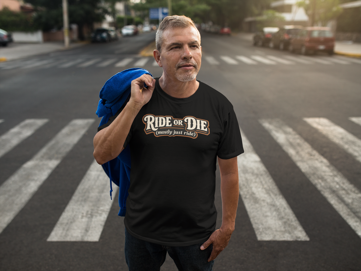 A man standing on a city crosswalk wearing a black graphic t-shirt with 'Ride or Die Mostly Just Ride' He has a blue jacket draped over his shoulder, with a blurred street background.