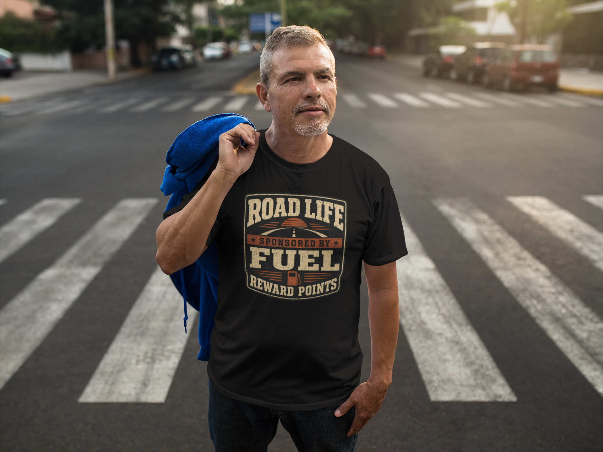 A man standing on a city crosswalk wearing a black graphic t-shirt with a 'Road Life Sponsored By Fuel Reward Points' design, holding a blue jacket over his shoulder.
