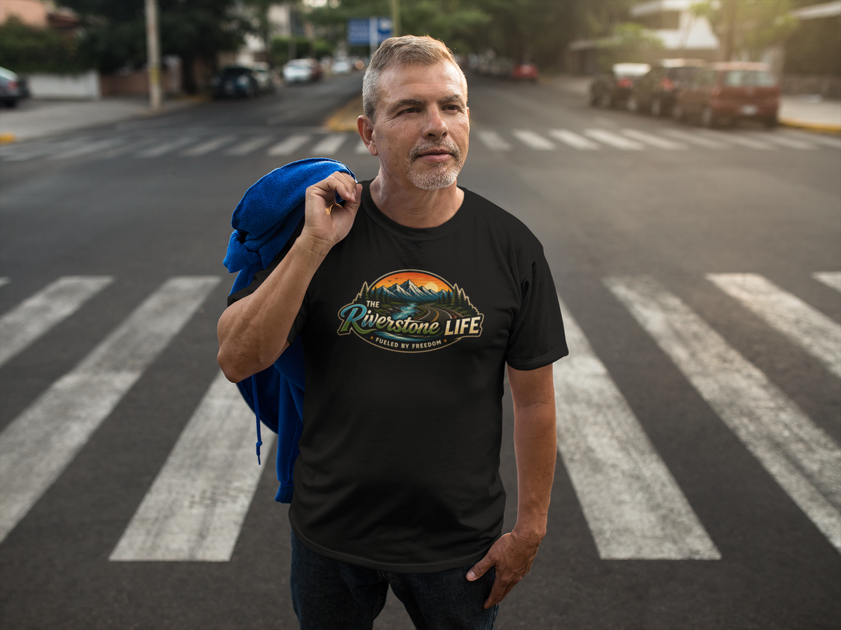 A man standing on a city crosswalk wearing a black graphic t-shirt with a colorful mountain landscape print that reads 'The Riverside Life' - 'Fueled by Freedom'. He has a blue jacket draped over his shoulder, with a blurred street background.