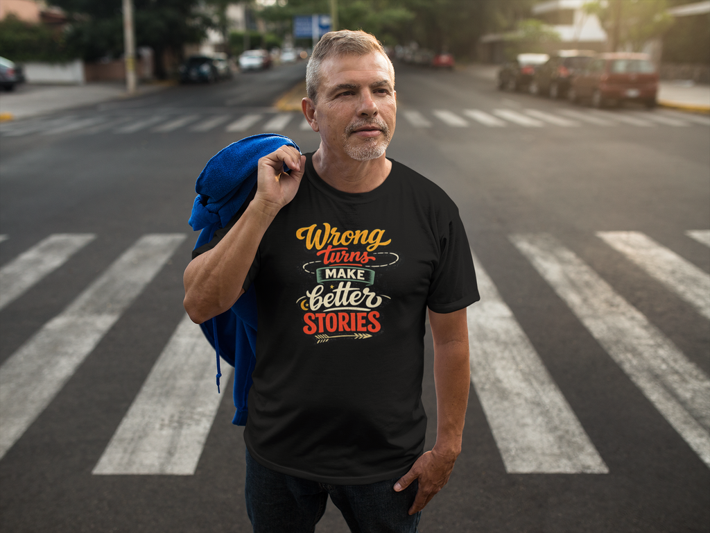 A man standing on a city crosswalk wearing a black graphic t-shirt with the text 'Wrong turns make Better Stories' in yellow, orange, and white lettering. He has a blue jacket draped over his shoulder, with a blurred street background.