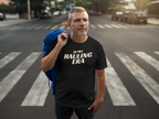 A man standing on a city crosswalk wearing a black graphic t-shirt that reads 'IN MY HAULING ERA' in bold white letters, with a blue jacket slung over his shoulder.