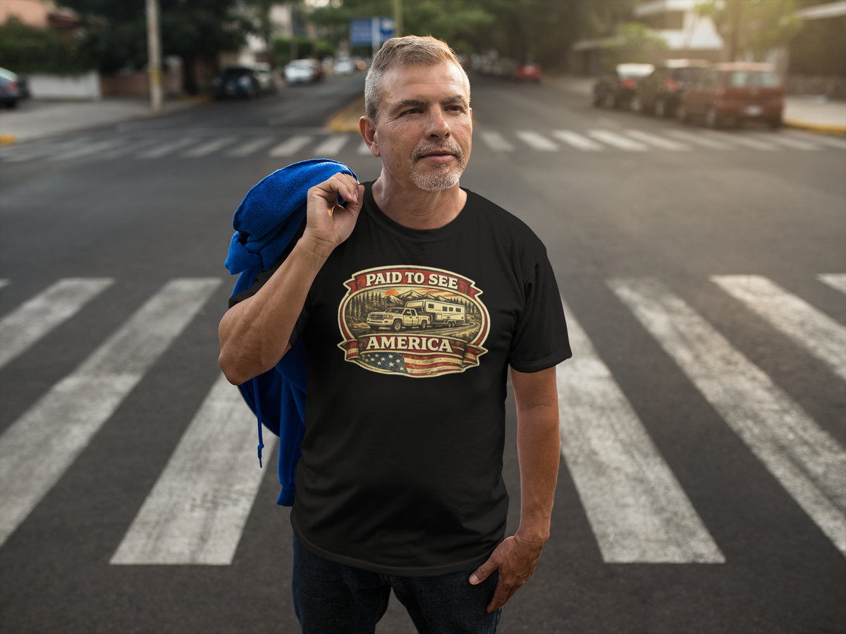 A man standing on a city crosswalk wearing a black graphic t-shirt with a vintage-style 'Paid to See America' travel poster design. He has a blue jacket draped over his shoulder, with a blurred street background.