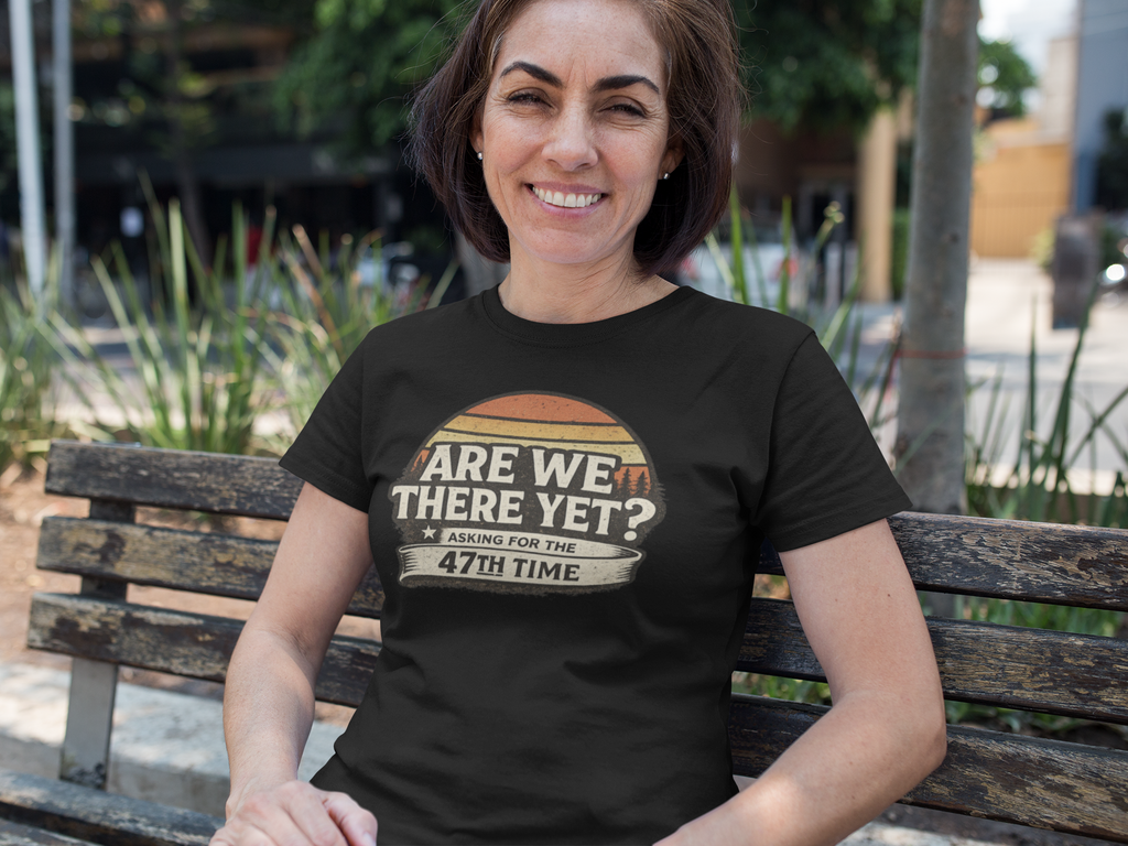 A woman sitting on a wooden bench outdoors wearing a black graphic t-shirt with a retro basketball design and the text 'ARE WE THERE YET? WAITING FOR THE 47TH TIME'.