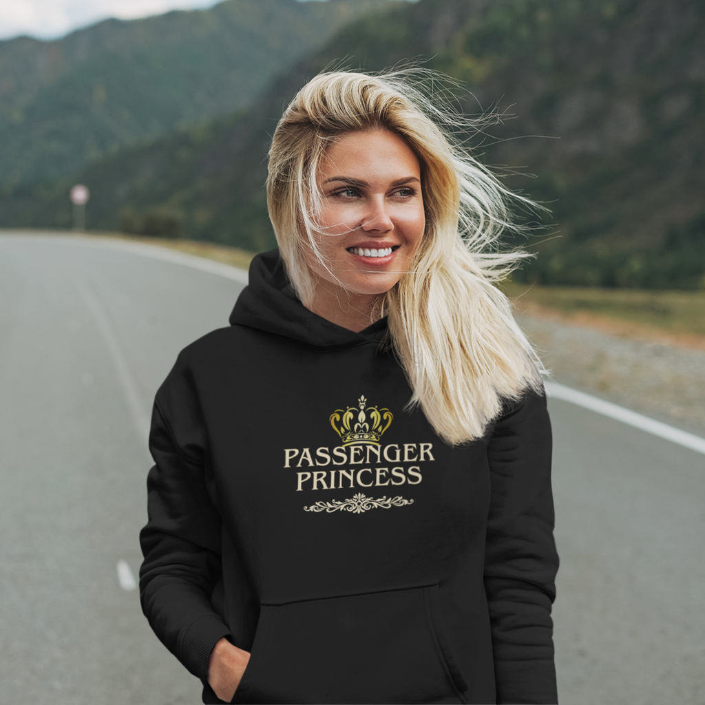 A woman with blonde hair stands on a road with mountains in the background, wearing a black hoodie with a gold crown graphic and the text 'Passenger Princess' on the chest.