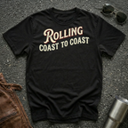 A black short-sleeve graphic T-shirt laid flat on a dark textured surface, with sunglasses, brown leather jacket, and a metal tumbler nearby. The shirt has large distressed text reading 'Rolling Coast to Coast' across the chest.