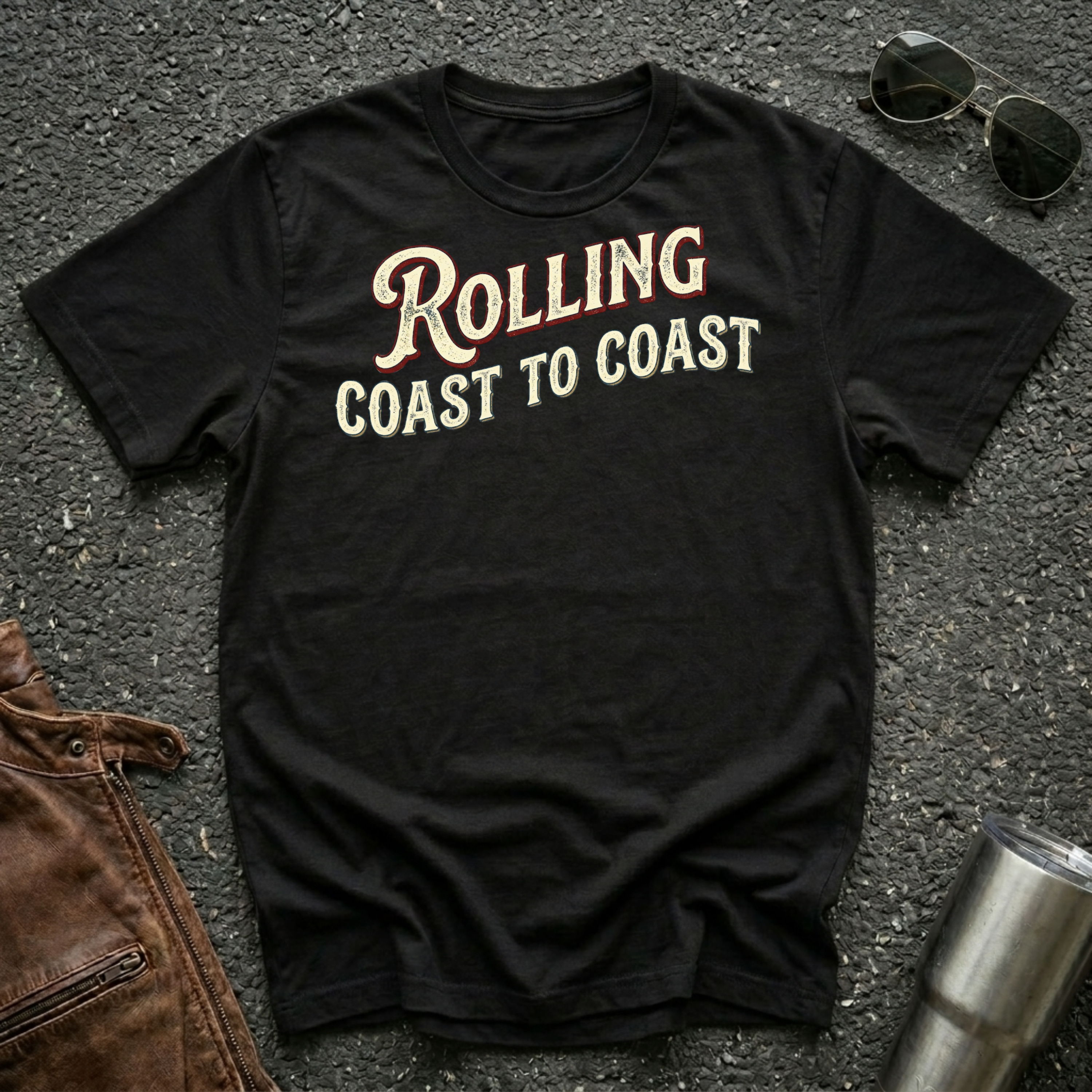 A black short-sleeve graphic T-shirt laid flat on a dark textured surface, with sunglasses, brown leather jacket, and a metal tumbler nearby. The shirt has large distressed text reading 'Rolling Coast to Coast' across the chest.