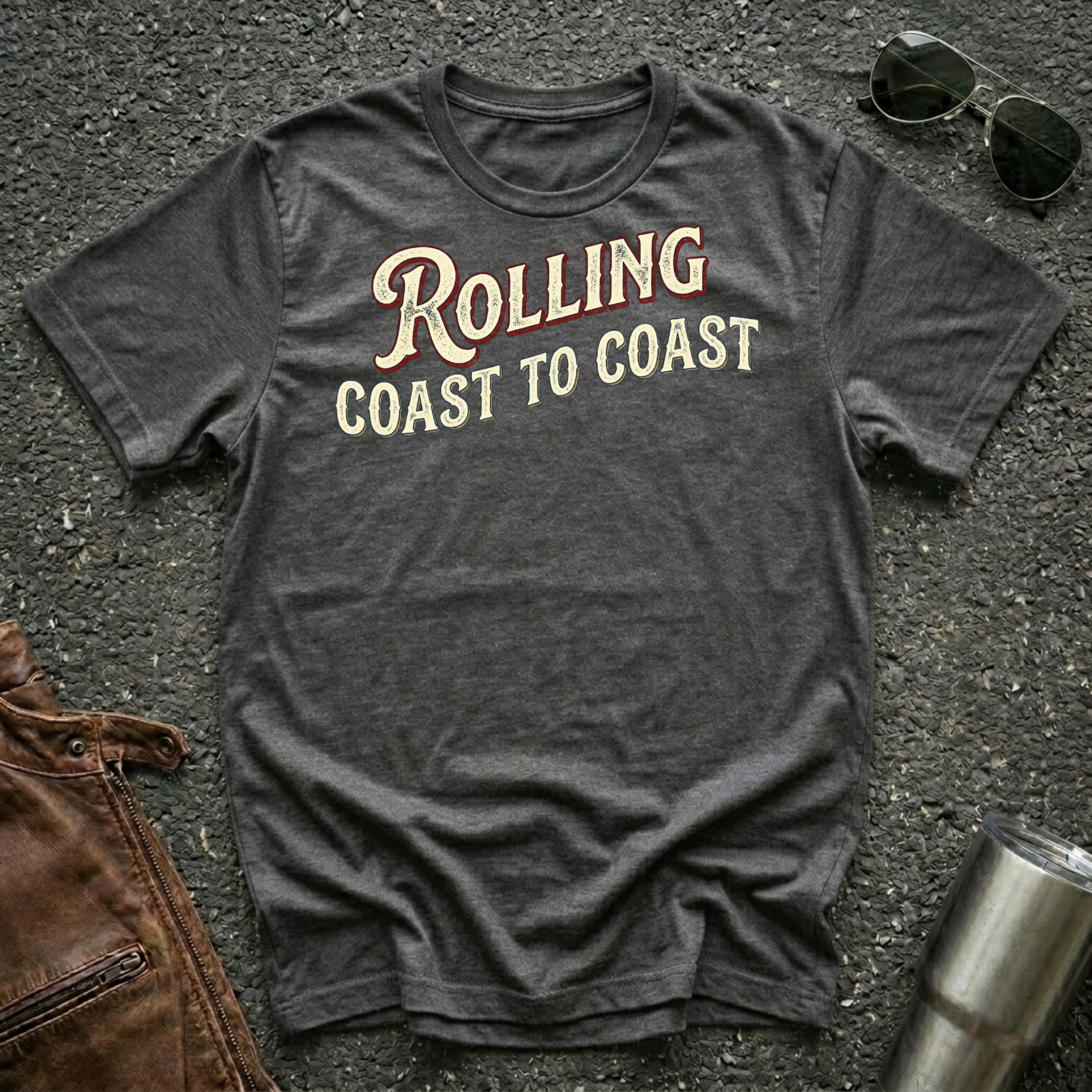 A dark heather short-sleeve graphic T-shirt laid flat on a dark textured surface, with sunglasses, brown leather jacket, and a metal tumbler nearby. The shirt has large distressed text reading 'Rolling Coast to Coast' across the chest.