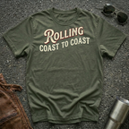 A military green short-sleeve graphic T-shirt laid flat on a dark textured surface, with sunglasses, brown leather jacket, and a metal tumbler nearby. The shirt has large distressed text reading 'Rolling Coast to Coast' across the chest.