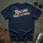 A navy short-sleeve graphic T-shirt laid flat on a dark textured surface, with sunglasses, brown leather jacket, and a metal tumbler nearby. The shirt has large distressed text reading 'Rolling Coast to Coast' across the chest.