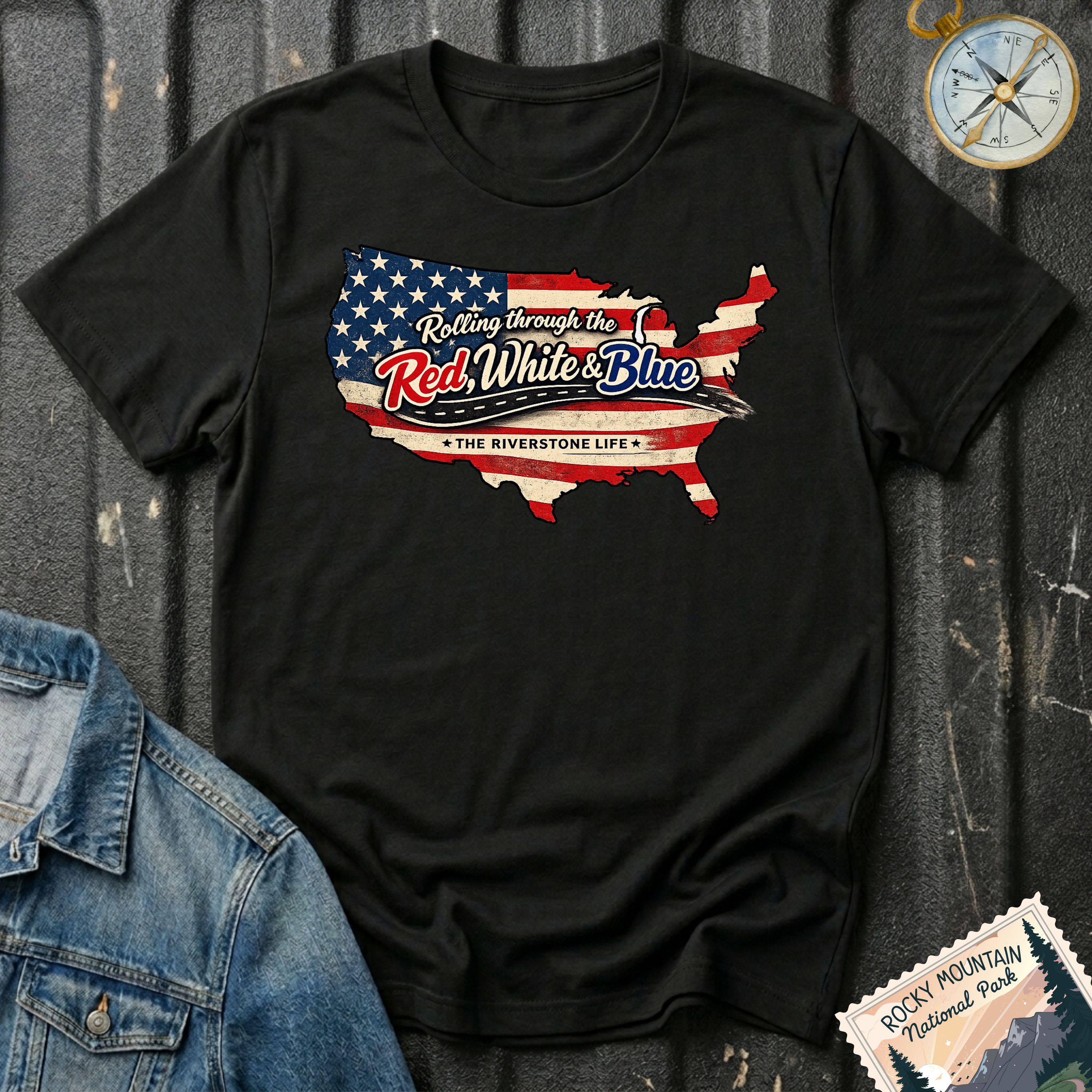 A black short-sleeve graphic T-shirt laid flat on a dark textured surface, featuring a USA map silhouette filled with an American flag design and the text 'Rolling through the Red, White & Blue' and 'The Riverstone Life.' A denim jacket corner, compass, and a 'Rocky Mountain National Park' stamp-style graphic are placed around it as styling props.
