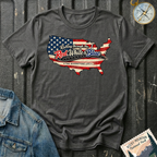 A dark heather graphic t-shirt laid flat on a dark textured background, styled with a denim jacket corner and a compass. The t-shirt features a graphic of the United States map filled with the American flag pattern and the text 'Rolling through the Red, White & Blue' and 'THE RIVERSTONE LIFE'.