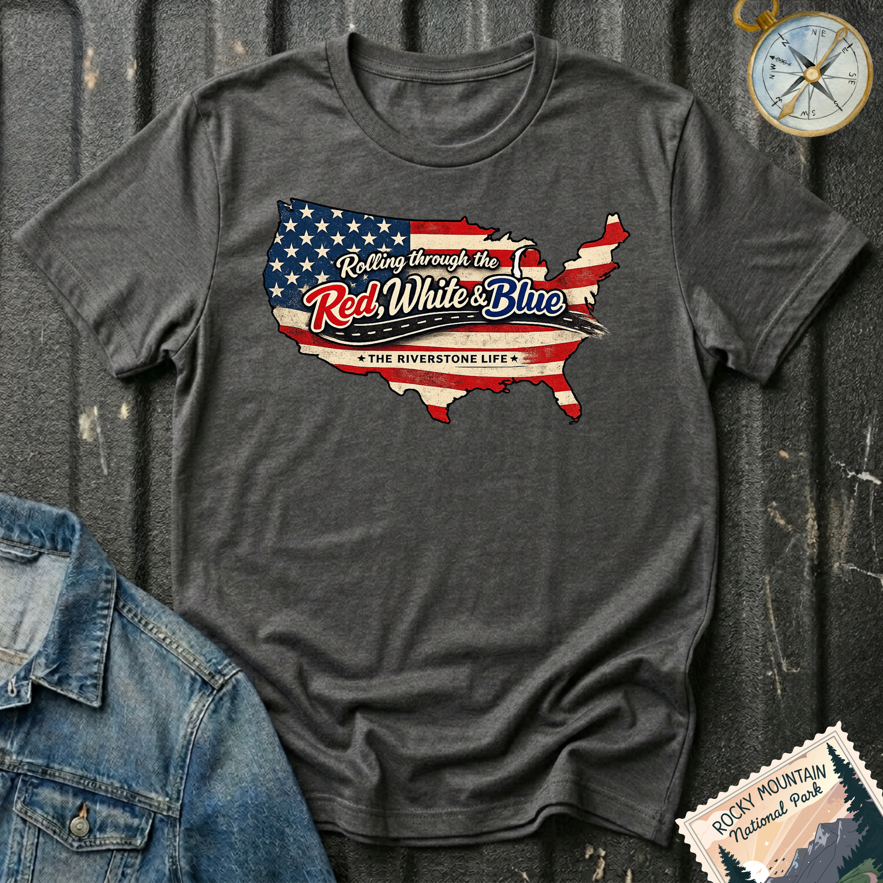 A dark heather graphic t-shirt laid flat on a dark textured background, styled with a denim jacket corner and a compass. The t-shirt features a graphic of the United States map filled with the American flag pattern and the text 'Rolling through the Red, White & Blue' and 'THE RIVERSTONE LIFE'.