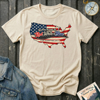 A beige graphic t-shirt laid flat on a dark wood background, styled with a denim jacket corner and a compass. The t-shirt features a map of the United States filled with the American flag pattern and the text 'Rolling through the Red, White & Blue' and 'THE RIVERSTONE LIFE'.