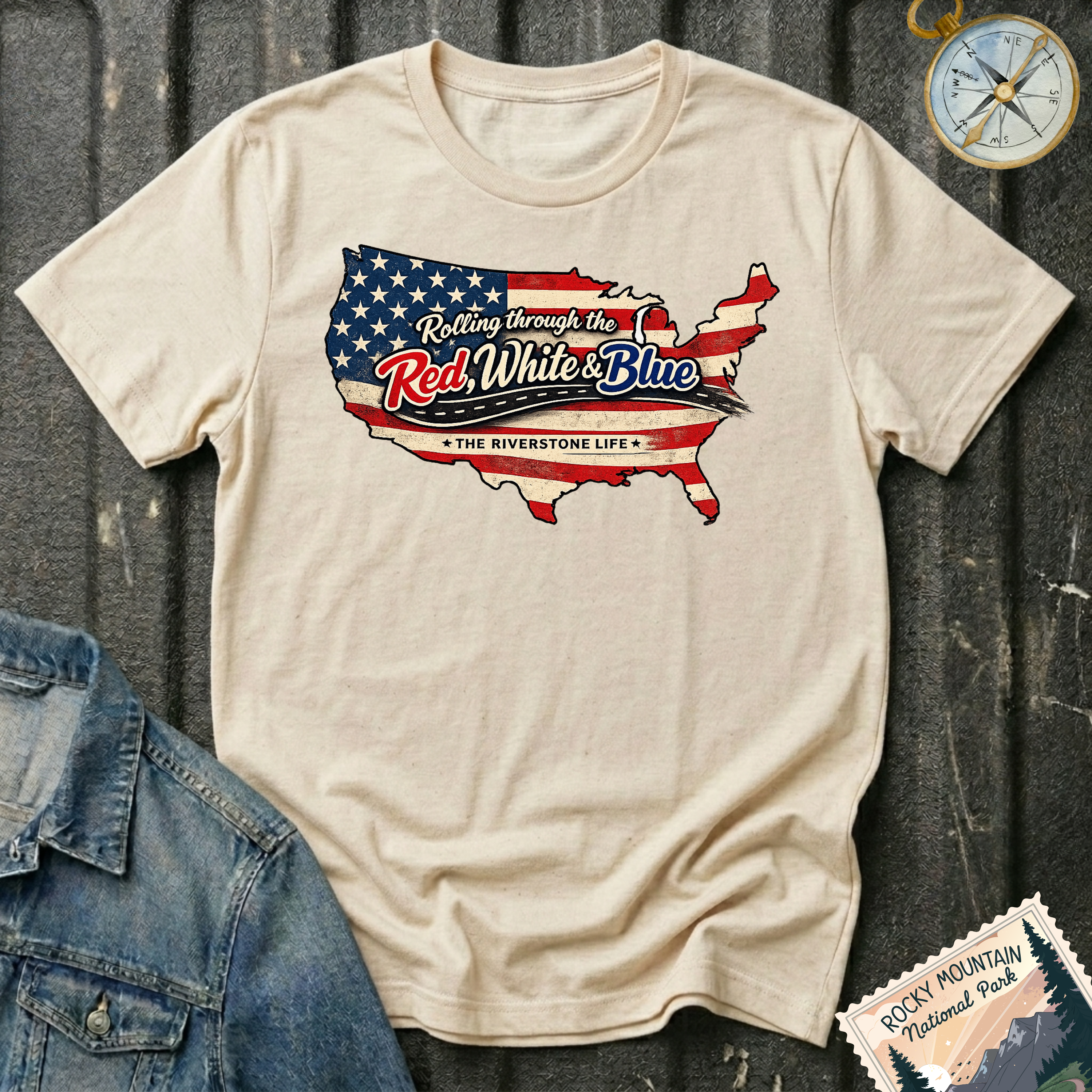 A beige graphic t-shirt laid flat on a dark wood background, styled with a denim jacket corner and a compass. The t-shirt features a map of the United States filled with the American flag pattern and the text 'Rolling through the Red, White & Blue' and 'THE RIVERSTONE LIFE'.