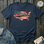 A navy short-sleeve t-shirt laid flat on a dark textured background, featuring a graphic of the United States map filled with an American flag pattern and the text 'Rolling through the Red, White & Blue' and 'THE RIVERSTONE LIFE'. A compass and denim jacket are styled around it.