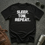 A black short-sleeve graphic T-shirt laid flat on a dark asphalt-like surface, with white grayish distressed text reading “SLEEP, TOW, REPEAT.” Sunglasses, brown leather jacket and a metal tumbler are placed around it as styling props.
