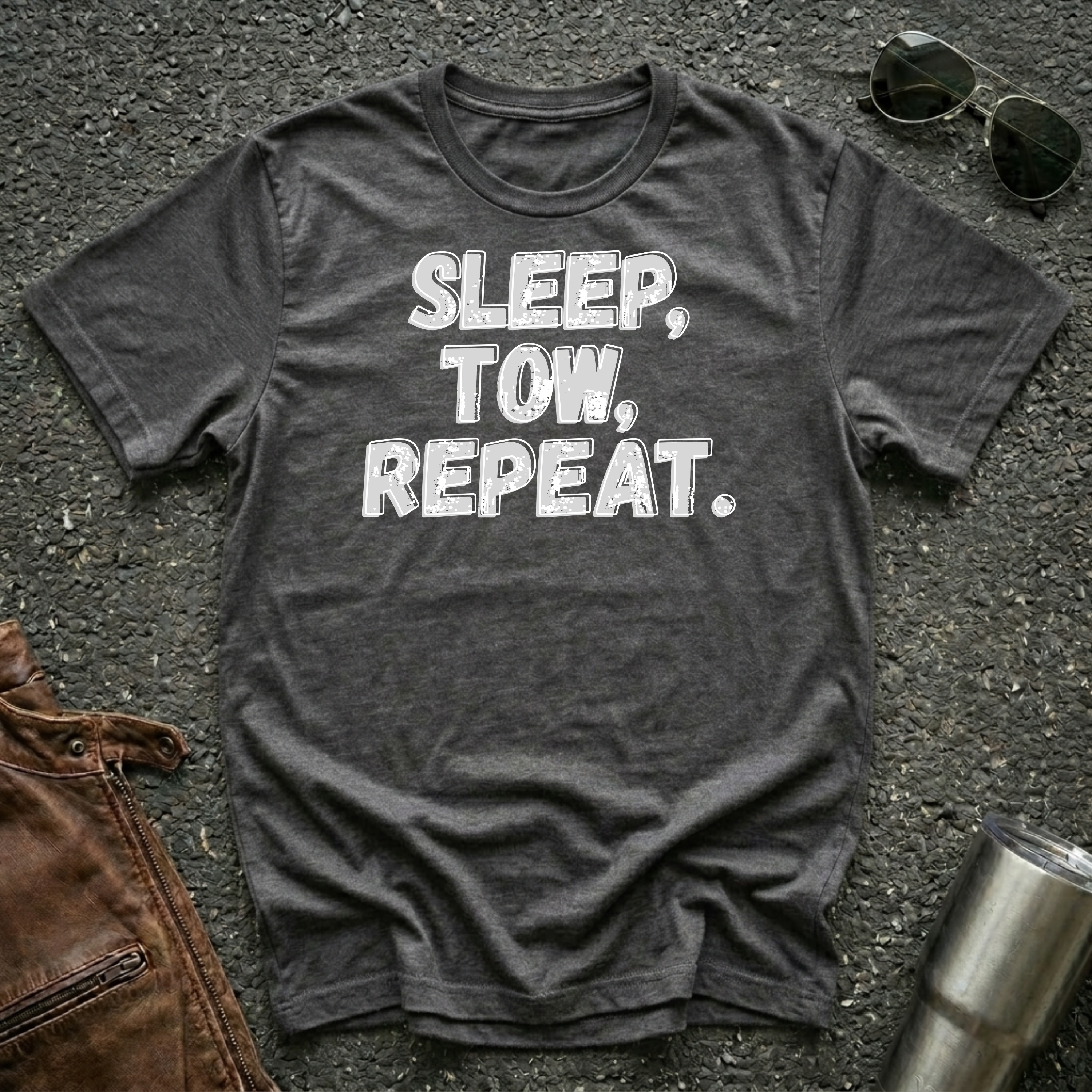 A dark heather gray graphic t-shirt with the text 'SLEEP, TOW, REPEAT.' printed in white gray-ish distressed letters, laid flat on a dark asphalt surface with sunglasses, a brown leather jacket, and a metal tumbler as props.