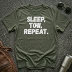 An olive green crewneck t-shirt with the text 'SLEEP, TOW, REPEAT.' printed in white gray-ish distressed letters, laid flat on a dark asphalt surface with sunglasses, a brown leather jacket, and a metal tumbler nearby.