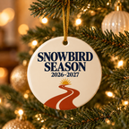 A round white ceramic Christmas ornament hanging from a gold string on a decorated Christmas tree. The ornament features the text 'SNOWBIRD SEASON 2026-2027' in dark blue, with a stylized red road and yellow sun graphic below.