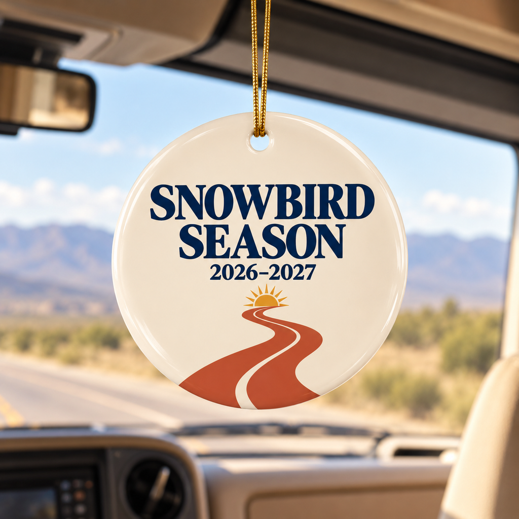 A round ceramic snowbird season ornament hanging from a car windshield. The ornament is cream-colored with navy blue text reading 'SNOWBIRD SEASON 2026-2027' and a graphic of a winding orange road leading to a yellow sun.