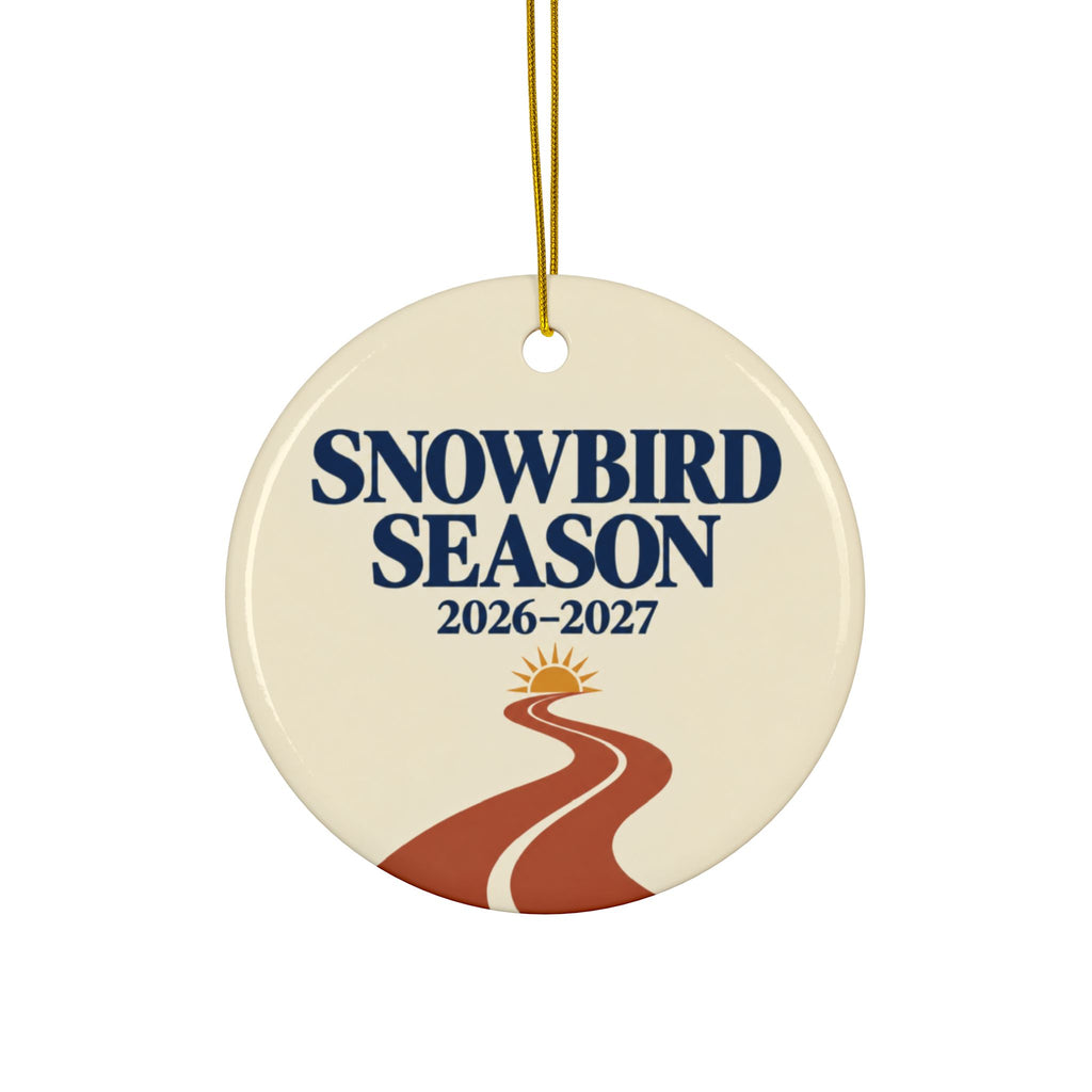 A round ceramic ornament hanging from a gold string, featuring the text 'SNOWBIRD SEASON 2026-2027' above a stylized winding road and sun graphic on an off-white background.