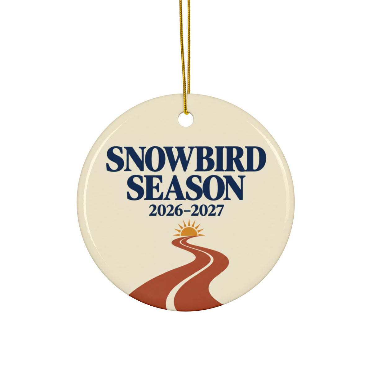 A round ceramic ornament hanging from a gold string, featuring the text 'SNOWBIRD SEASON 2026-2027' above a stylized winding road and sun graphic on an off-white background.