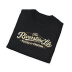 A black folded t-shirt with a vintage-style cream and gold graphic reading 'The Riverstone Life FUELED BY FREEDOM' on a clean white background.